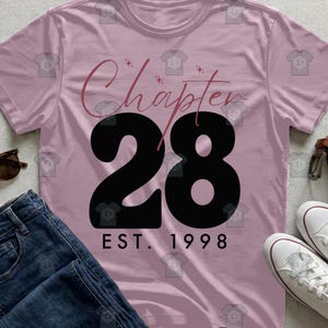 May include: A light pink t-shirt with the words "Chapter 28" in a cursive font and the number "28" in bold black. Below the number is "EST. 1998" in black. The shirt is paired with blue jeans and white sneakers.