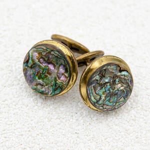 May include: A pair of round cufflinks featuring iridescent abalone shell inlays set within polished brass frames. The shell displays a swirling pattern of green, purple, and blue tones, while the metal edges have a warm, metallic gold-toned finish.