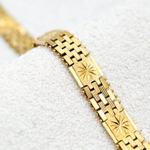 May include: A gold-colored bracelet with a starburst design on rectangular links. The bracelet is made of a shiny, metallic material and is displayed on a white, textured surface. The bracelet's design features a series of rectangular links.