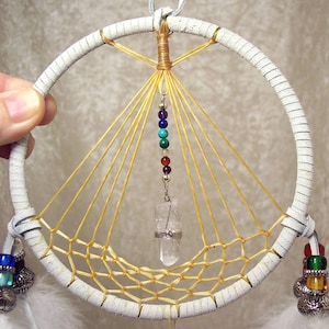 May include: White leather dreamcatcher with a yellow macrame web and a crystal pendant. The dreamcatcher has a beaded string with seven colorful beads hanging from the center.