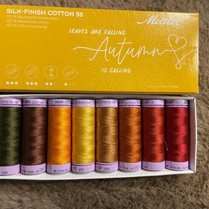 Könnte beinhalten: Ein Mettler Silk-Finish Cotton 50 Garnset in einer gelben Schachtel mit der Aufschrift Leaves Are Falling Autumn Is Calling. Die Schachtel enthält acht Garnrollen in den Farben Olivgrün, Braun, Orange, Gelb, Bronze, Kupfer, Rost und Dunkelrot.
