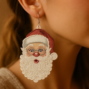 Op de afbeelding: Geborduurde kerstman oorbellen. De oorbellen hebben een rode hoed, witte baard en ronde bril. Het gezicht van de kerstman is gedetailleerd met een rode neus en roze wangen. De oorbellen zijn bevestigd aan een zilverkleurige haak.