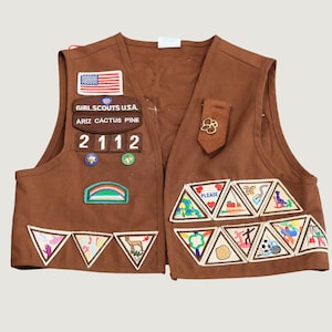 Op de afbeelding: Een bruin Girl Scouts U.S.A. vest met een Amerikaanse vlag-patch, een Ariz Cactus Pine council-patch, het nummer 2112, twee ster-speldjes, een regenboog-patch en talloze driehoekige geborduurde badges met diverse activiteiten en symbolen.