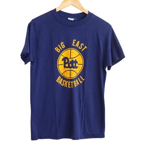 May include: Navy blue t-shirt with a yellow basketball graphic. The graphic includes the text "BIG EAST", "Pitt", and "BASKETBALL". The shirt is hanging on a wooden hanger against a white background.
