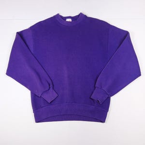 May include: A solid purple long-sleeve crewneck sweatshirt with ribbed cuffs and a ribbed hem. The garment features a simple, plain design with a small white brand label visible at the inside of the neckline collar, providing a clean look.