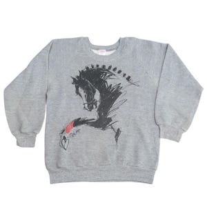 Op de afbeelding: Een grijs gemêleerd sweatshirt met lange mouwen en een zwarte schetsachtige illustratie van een paardenkop en -hals. Het paard heeft een rode band om het voorbeen en een klein hartje onderaan de afbeelding, met een ronde halslijn.