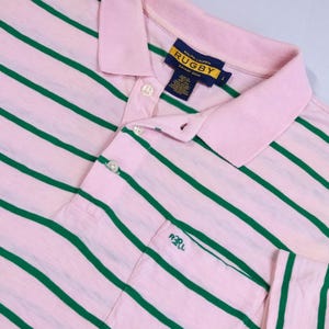 May include: Pink and green striped rugby shirt with a collared neckline and button-up placket. The shirt features a small pocket on the chest and a label that reads "RALPH LAUREN RUGBY EST. 2004".