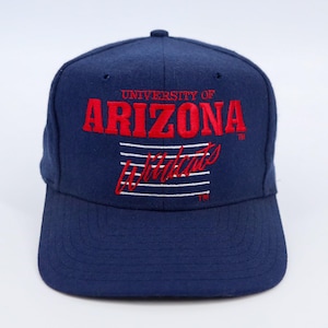 May include: A navy blue baseball cap with the words "UNIVERSITY OF ARIZONA" in red block letters. Below, the word "Wildcats" is in red script, with white horizontal lines. The cap has a curved brim.