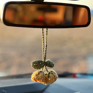 May include: A handmade car charm hanging from a rearview mirror. The charm features two yellow and green crocheted flowers with a green crocheted leaf. The mirror reflects a blurred outdoor scene.