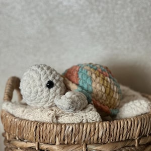 May include: A handmade crochet turtle toy with a cream-colored head and limbs, and a multicolored shell in shades of peach, blue, and yellow. The turtle rests in a woven basket lined with a cream-colored blanket, creating a cozy scene.