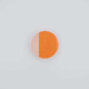May include: A round, two-toned button with a split design. One half is a light peach color, and the other is a vibrant orange. The button is set against a plain white background, highlighting its simple, yet striking design.