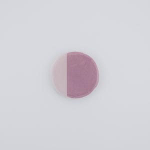May include: A circular object divided into two vertical halves. One side is a pale pink, the other a muted purple. The item is placed on a plain white backdrop, giving a clean, minimalist look. The texture seems soft and slightly porous.