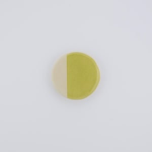 May include: A round, two-toned ceramic coaster. One half is a creamy off-white, and the other is a muted, yellowish-green. The coaster has a slightly irregular, organic shape, and is set against a plain white background.