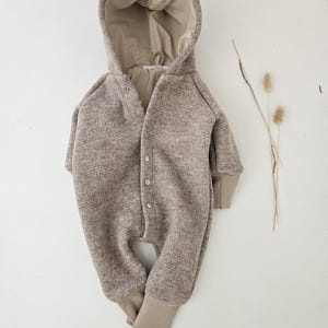 Könnte beinhalten: Ein neutralfarbener Baby-Overall mit Kapuze. Das einteilige Kleidungsstück hat eine Knopfleiste vorne, lange Ärmel und gerippte Bündchen an Ärmeln und Knöcheln. Der Kapuzen-Overall ist aus einem weichen, strukturierten Stoff gefertigt.