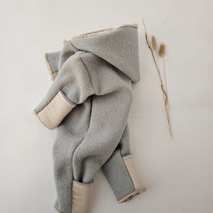 Peut inclure: Combinaison à capuche gris clair avec des empiècements beiges aux poignets et à l'intérieur de la capuche. Le tissu doux, semblable à de la laine, est plié sur une surface blanche. Quelques tiges séchées sont placées à droite.