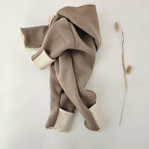 Könnte beinhalten: Ein brauner Kapuzen-Overall mit cremefarbenen Zierleisten. Der weiche, wollähnliche Stoff ist drapiert und zeigt das Design des Kleidungsstücks. Die Manschetten und das Kapuzenfutter sind in einem kontrastierenden Cremeton gehalten. Ein Zweig trockenes Gras setzt einen natürlichen Akzent.