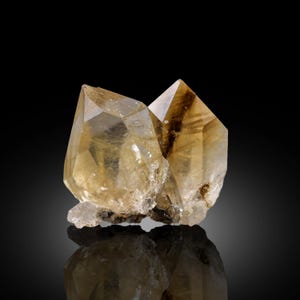 Puede incluir: Dos cristales de cuarzo de color amarillo dorado con puntas afiladas. Los cristales son translúcidos, con estrías e inclusiones internas visibles. La base de los cristales es rugosa, blanquecina, sobre un fondo negro.