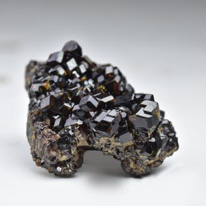 May include: A cluster of dark brown, lustrous garnet crystals. The crystals have a cubic shape and are densely packed together, creating a textured surface. The mineral specimen sits on a rough, earthy matrix.