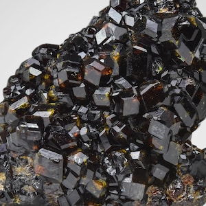 May include: Close-up of a cluster of dark, lustrous crystals. The crystals are primarily black with hints of amber and brown, showcasing a complex, geometric structure. The overall texture is rough and uneven, with sharp edges and reflective surfaces.