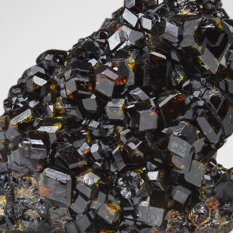 May include: Close-up of a cluster of dark, lustrous crystals. The crystals are primarily black with hints of amber and brown, showcasing a complex, geometric structure. The overall texture is rough and uneven, with sharp edges and reflective surfaces.