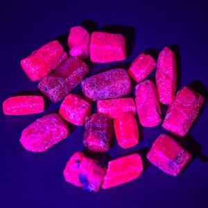 May include: A collection of small, rough-cut, raw rubies scattered across a dark surface. The stones exhibit a vibrant, intense neon pink and red glow, characteristic of fluorescence when exposed to ultraviolet light, showing irregular shapes.