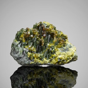 Op de afbeelding: Een close-up van een mineraal specimen met een cluster van groene, naaldachtige kristallen en geelbruine accenten. Het mineraal ligt op een grijze, getextureerde basis, met zijn weerspiegeling eronder.