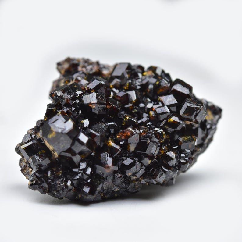May include: A cluster of lustrous, dark brown garnet crystals. The crystals have a geometric, cubic shape and are densely packed together. The surface of the crystals reflects light, creating a sparkling effect. The mineral specimen is set against a white background.