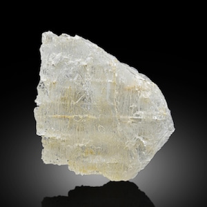 Può includere: Un esemplare di minerale traslucido e cristallino con una forma ruvida e irregolare. La pietra ha un colore bianco sporco chiaro con sottili inclusioni gialle e marroni. La superficie ha un aspetto strutturato, con strutture e linee interne visibili.