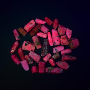 Puede incluir: Un conjunto de piedras preciosas de rubí en bruto, sin cortar, en tonos rojos y rosas, dispuestas sobre una superficie negra. Las piedras tienen una textura natural y rugosa, con variaciones de color y forma.