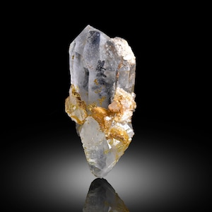 Puede incluir: Un cristal de cuarzo transparente con una punta y una superficie rugosa y texturizada. El cristal tiene inclusiones y está acentuado con parches de depósitos minerales de color marrón dorado. El cristal se muestra sobre un fondo negro.