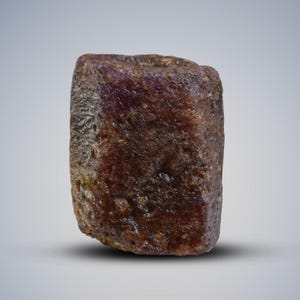 Pode incluir: Uma gema de rubi natural, em bruto e não polida. O rubi tem uma cor castanho-avermelhada escura e uma superfície texturizada. A gema é de forma retangular, mostrando sua forma bruta e natural. Adequado para a fabricação de joias ou colecionadores de gemas. Mede aproximadamente 2.5 cm.