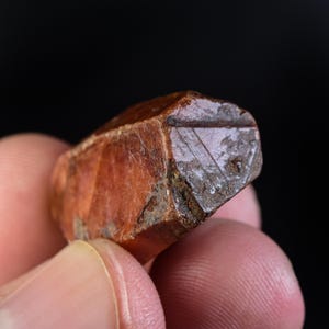 May include: A close-up of a rough, reddish-brown mineral specimen. The stone has a faceted, geometric shape with a triangular pattern carved into one end. The mineral is held between two fingers against a black background.
