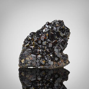 May include: A cluster of lustrous, dark brown and black crystals. The crystals have a cubic shape and are densely packed together, creating a rough, textured surface. The mineral formation is set against a neutral background, with a reflective surface below.