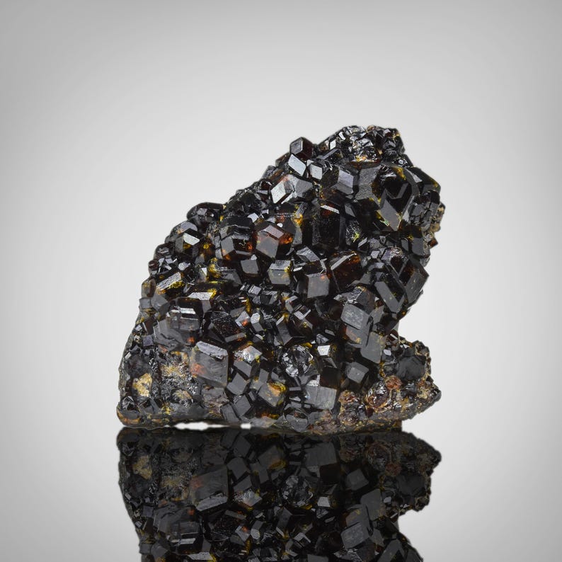 May include: A cluster of lustrous, dark brown and black crystals. The crystals have a cubic shape and are densely packed together, creating a rough, textured surface. The mineral formation is set against a neutral background, with a reflective surface below.