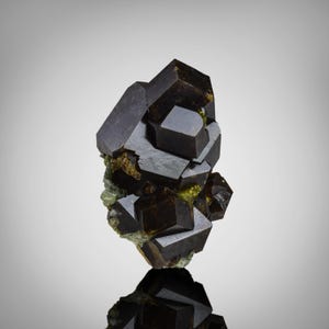 Pode incluir: Um grupo de minerais castanhos escuros com formações cristalinas geométricas. Os cristais têm uma superfície refletora e são colocados sobre um fundo claro. A base do grupo tem um tom verde e amarelo.
