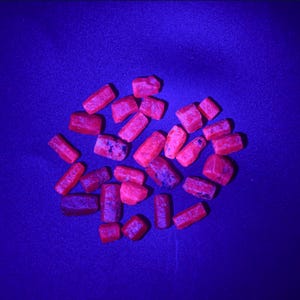 Pode incluir: Uma coleção de pedras preciosas vermelhas em bruto espalhadas sobre uma superfície azul escura. As pedras variam em tamanho e forma, com algumas mostrando inclusões mais escuras, arroxeadas. A iluminação dá às pedras uma aparência vibrante e brilhante.