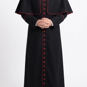 May include: A black cassock with red trim and buttons. The garment features a capelet and is worn with a white collared shirt. The buttons run down the front of the cassock. The person is wearing black shoes.