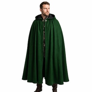 May include: A floor-length forest green cloak featuring a deep hood and a circular silver-toned metal clasp at the neckline. The garment is draped over a dark brown leather-like tunic, showcasing a full, pleated silhouette that falls to the ankles.