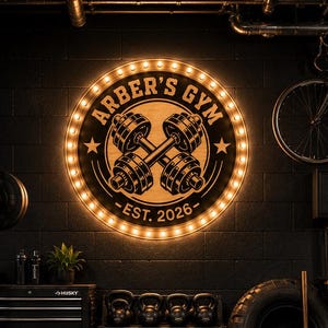 May include: A gym interior with a large, circular sign that reads "Arber's Gym" with a dumbbell graphic and "Est. 2026". The sign is illuminated with warm lights. The room features weightlifting equipment, including barbells, kettlebells, and a tire.