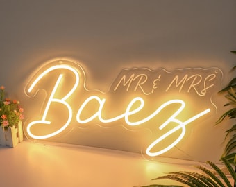 May include: A warm white neon sign displays the name "BaeZ" in a flowing script, with "MR & MRS" above. The sign is mounted on a clear backing, suitable for wedding decorations or home interiors.