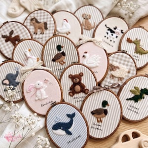 Könnte beinhalten: Eine Sammlung ovaler Holzstickrahmen mit Filz-Tierapplikationen auf Stoffhintergründen. Jeder Rahmen zeigt ein einzigartiges Tier wie einen Bären, Wal, eine Ente oder einen Hasen, wobei die Namen unter den Figuren eingestickt sind.