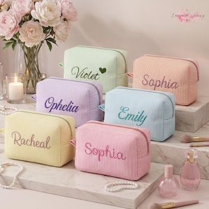 Op de afbeelding: Zes rechthoekige stoffen make-uptasjes met verticale pastelkleurige strepen, elk voorzien van een geborduurde naam. De tasjes zijn lichtgroen, oranje, paars, lichtblauw, geel en roze, met ritsen en bijpassend garen in de borduursels.