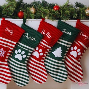 Pode incluir: Meias de Natal personalizadas em vermelho e verde, com detalhes em branco. Cada meia é bordada com um nome em branco. Algumas meias apresentam pegadas de patas ou um desenho de árvore de Natal.
