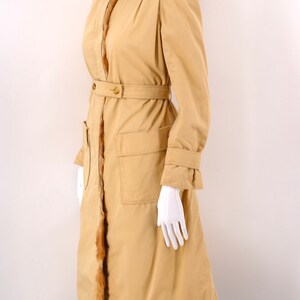 May include: A long, tan coat with a fur-trimmed hood and a belt. The coat has two large pockets on the front and a fur trim along the sides.