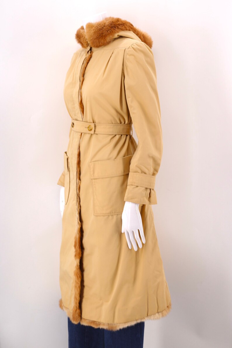 May include: A long, tan coat with a fur-trimmed hood and a belt. The coat has two large pockets on the front and a fur trim along the sides.