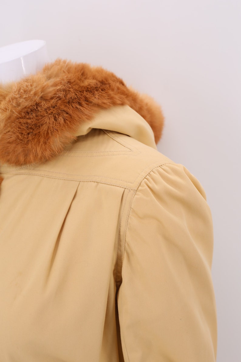 May include: A beige hooded jacket with a brown fur collar. The jacket has a long sleeve and a fitted silhouette.