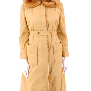 May include: A long, tan coat with a fur-lined hood and a belt. The coat has two large pockets on the front and a fur trim along the bottom.