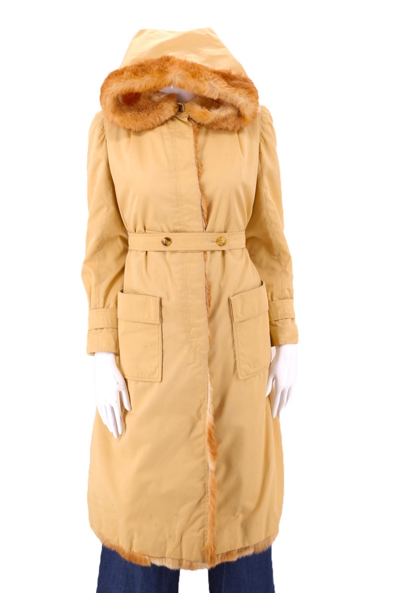 May include: A long, tan coat with a fur-lined hood and a belt. The coat has two large pockets on the front and a fur trim along the bottom.
