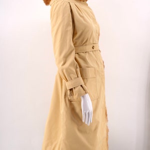 May include: A long, beige coat with a fur-trimmed hood and a belt. The coat has a large pocket on the right side and a fur trim along the bottom.