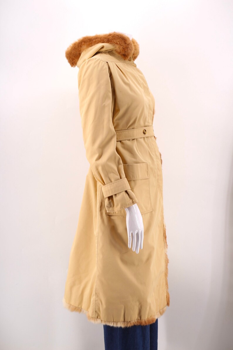 May include: A long, beige coat with a fur-trimmed hood and a belt. The coat has a large pocket on the right side and a fur trim along the bottom.